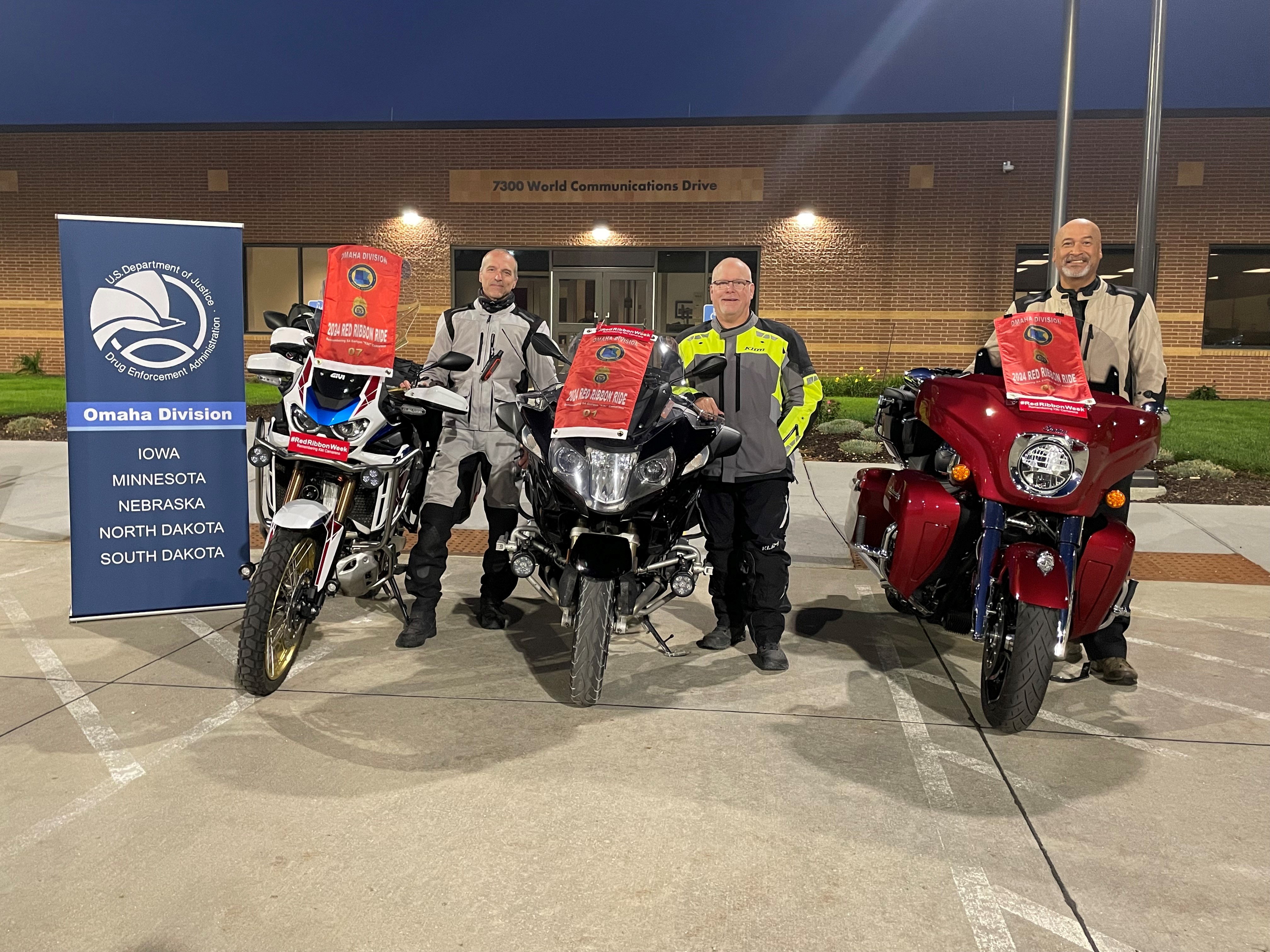 Three members of DEA stand next to their motorcycles and a blue DEA banner.